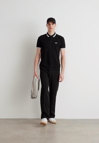 Schwarzes Polohemd mit weißen Kragenakzenten, kombiniert mit schwarzen Hosen und weißen Sneakers. Das Model hält einen hellgrauen Sweatshirt.