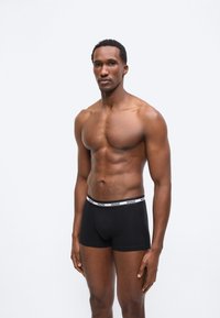 Μυώδης άνδρας στέκεται μπροστά σε λευκό φόντο φορώντας μαύρα εσώρουχα boxer με το λογότυπο της Hugo, κοιτάζοντας ελαφρώς προς το πλάι με ουδέτερη έκφραση.