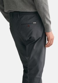 Dunkelgraue Hose aus Baumwollmischung, mit einer Gesäßtasche mit einem braunen Knopf und einem kleinen metallischen Logo-Akzent in der Nähe der Tasche.