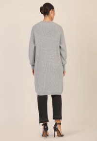 Lange mouwen, lichtgrijze gebreide cardigan met een geribbelde textuur, die onder de heupen valt, gecombineerd met cropped zwarte jeans en hoge hakken.