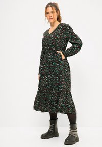 Vestido negro con un patrón de zigzag multicolor, mangas largas y escote en V. Presenta un dobladillo en capas y bolsillos laterales, combinado con botas negras gruesas.