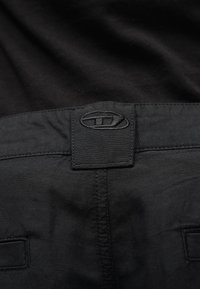 Μαύροι παντελόνες με ραμμένη ετικέτα στη μέση που φέρει ένα οβάλ λογότυπο διαιρεμένο οριζόντια, ραμμένο με κλωστή στο ίδιο μαύρο χρώμα.