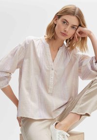 Blusa leggera di colore crema con sottili righe pastello, apertura a bottoni, scollo a V e maniche 3/4. Si abbina a pantaloni beige e sneakers bianche.