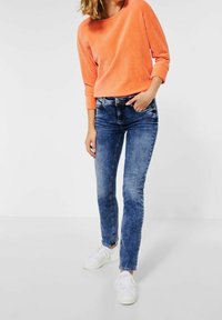 Orange ribbad tröja, blå slim-fit jeans och vita sneakers. Tröjan har en lös passform med rund halsringning; jeansen har en klassisk design.