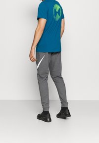 Camiseta color azul verdoso con un gráfico verde en la parte posterior, combinada con pantalones de chándal grises que tienen un logo blanco de Nike y zapatos deportivos negros.
