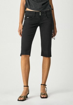 Pantalones capri de mezclilla negra con un diseño ajustado, dos bolsillos delanteros y dobladillos rematados, llevados con tacones negros con tiras.