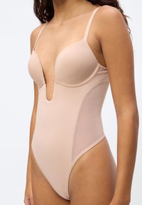 Γυναίκα που φοράει κορμάκι σε γυμνή απόχρωση με βαθύ V ντεκολτέ και λεπτές τιράντες, εφαρμοστή σχεδίαση και χωρίς ραφές στα cups, με φόντο απλό.