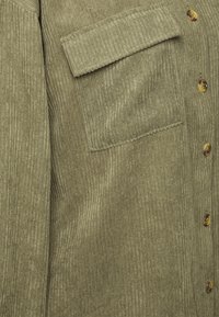Camisa de pana de color verde oliva con rayas verticales, que cuenta con un bolsillo en el pecho y cierres de botón marrón.