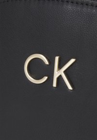 Schwarzes Lederprodukt mit einem goldenen "CK"-Logo, das eine glatte Textur mit Nahtdetails entlang der Ränder zeigt.