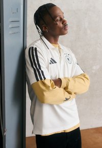 Polo branco com riscas pretas e brasão da Alemanha. Por baixo, uma camisa de mangas longas amarela. Calças pretas, de pé ao lado de um armário.