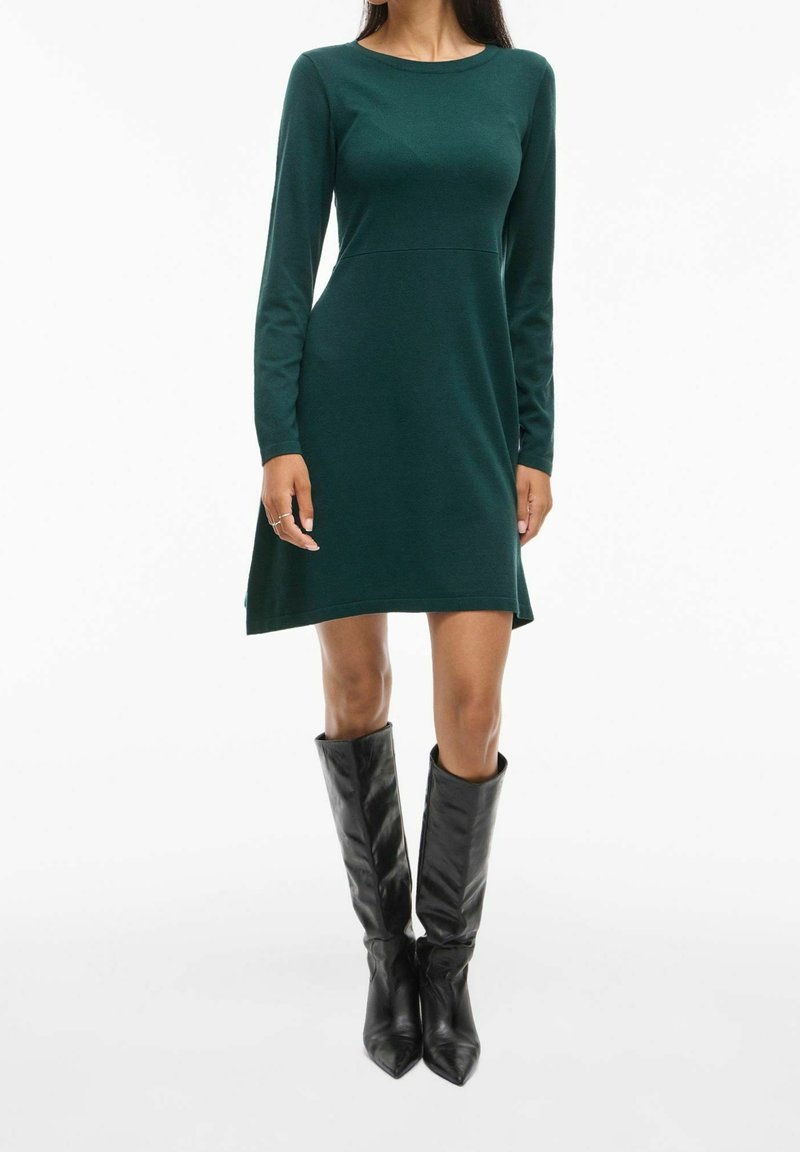 Femme portant une robe trapèze à manches longues de couleur vert foncé associée à des bottes en cuir noires montantes, posant sur un fond blanc.