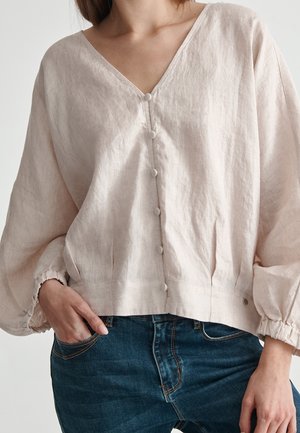 Vrouw draagt een losse lichtbeige V-halsblouse met stoffen bedekte knopen aan de voorkant en poflange mouwen, gecombineerd met een blauwe spijkerbroek.