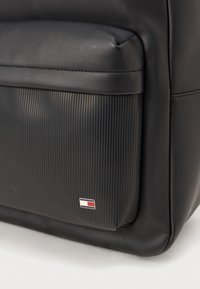 Čierna kožená taška s ryhovanou čiernou spodnou časťou, na ktorej sa nachádza malé logo v červenej, bielej a modrej farbe. Zaoblené okraje a prešívané detaily.