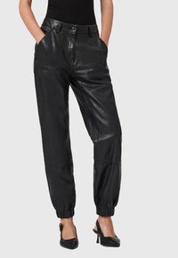 Vrouw in zwarte high-waisted leren joggerbroeken met elastische mouwen en zwarte puntige slingback hakken, met de handen in de zakken.