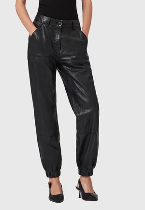 Donna che indossa pantaloni jogger in pelle neri a vita alta con polsini elastici e décolleté neri a punta con cinturino alla caviglia, mani in tasca.