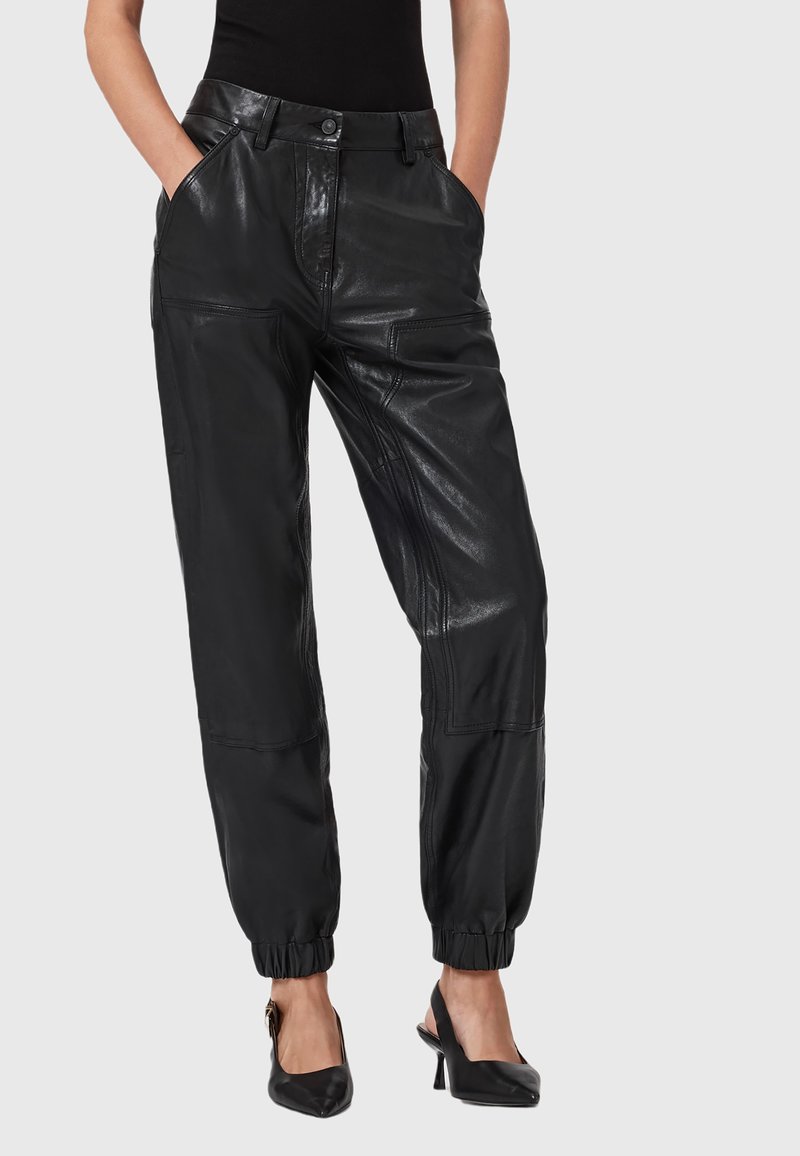 Vrouw in zwarte high-waisted leren joggerbroeken met elastische mouwen en zwarte puntige slingback hakken, met de handen in de zakken.