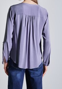 Blusa lunga a maniche con girocollo di colore viola chiaro, con dettagli arricciati sul retro e spacchi laterali, indossata sopra jeans blu scuro.