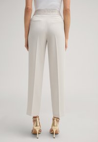 Helle beige weit geschnittene Hose mit einem strukturierten Taillenbund, der den Markennamen trägt, zwei Gesäßtaschen und einer glatten Stoffoberfläche.