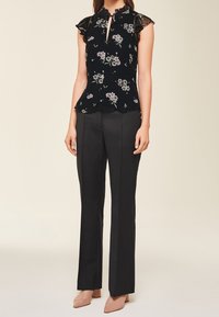 Kvinde iført sort blomstret ærmeløs bluse med flæser på skuldrene, sorte skræddersyede bukser og nude spidse hæle mod en beige baggrund.