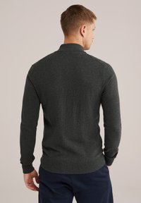 Rövid, világosbarna hajú férfi sötétszürke hosszúujjú pulóverben és tengerészkék nadrágban, háttal áll egy sima háttér előtt.