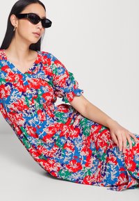 Vestido floral até ao joelho com decote em V, cintura franzida e mangas bufantes. O tecido apresenta um padrão vibrante de flores vermelhas, azuis e verdes.