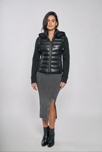 Kvinde iført sort, quiltet, lynlåset jakke med hætte, grå strikket nederdel med sideslids og sorte ankelstøvler mod en ensfarvet baggrund.