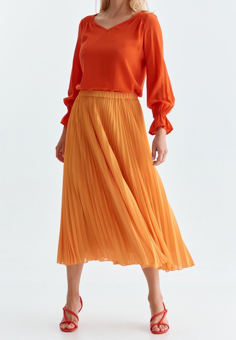 Femme portant une longue jupe plissée orange, un chemisier à manches longues orange vif et des sandales rouges à talons à brides, sur un fond uni.