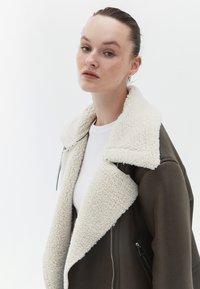 Brun läderjacka med en stor krämfärgad sherlingskrage och blixtlåsade sidofickor. Insidan är fodrad med ett mjukt, strukturerat material.