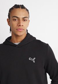 Felpa nera con cappuccio e cordino, dotata di un piccolo logo Puma bianco sul petto. Tessuto morbido, design aderente e superficie liscia.