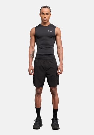Hombre con cabello trenzado que lleva una camiseta sin mangas ajustada negra y pantalones cortos cargo negros, de pie mirando hacia adelante sobre un fondo blanco.