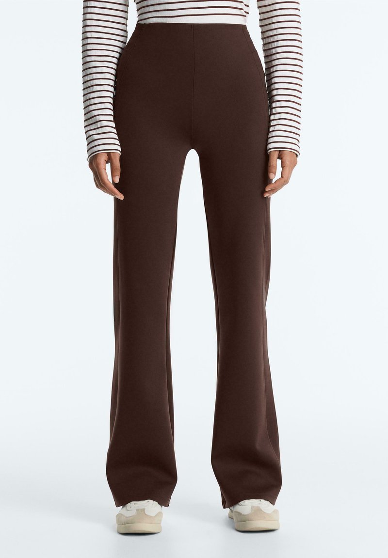 Person wearing high-waisted, wide-leg dark brown pants, striped long-sleeve shirt, and white sneakers, standing against white background.