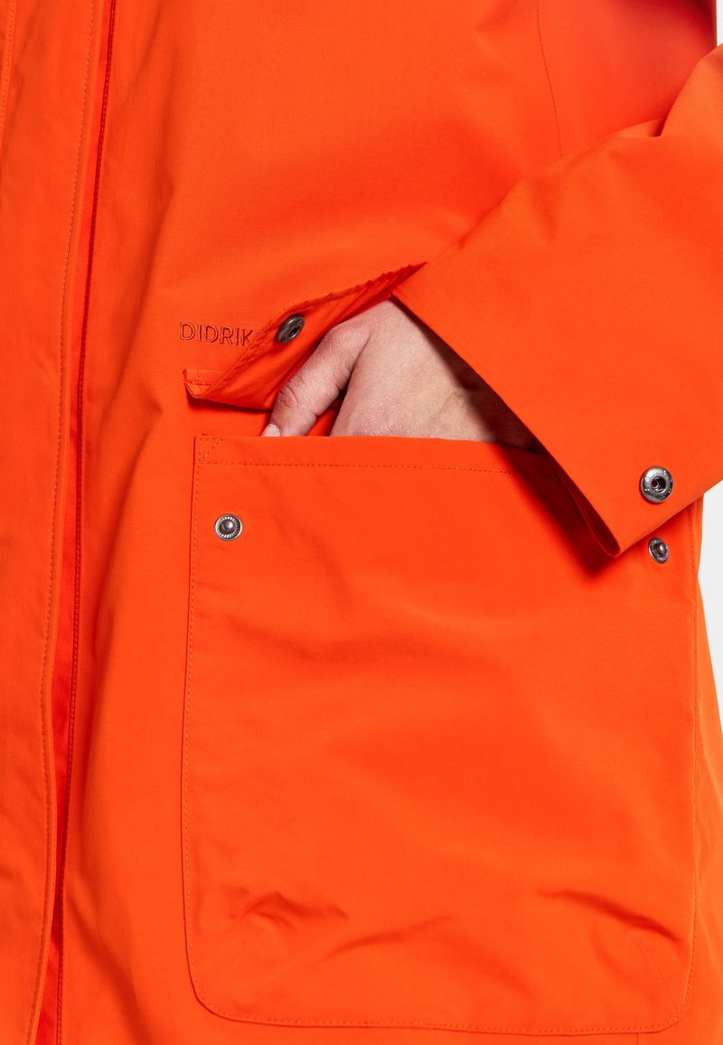 Orangene wasserdichte Jacke mit einer großen Seitentasche und Druckknopfverschluss, ausgestattet mit einer glatten Textur und minimalistischen Designdetails.