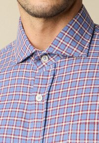 Primo piano del collo di un uomo e del colletto della camicia che mostrano una camicia a quadri blu e rossi con bottoni bianchi, parzialmente abbottonata.