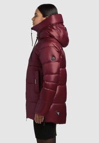 Kvinde i profil iført en skinnende maroon polstret jakke med høj krave og hætte, stående mod en ensfarvet lys baggrund.