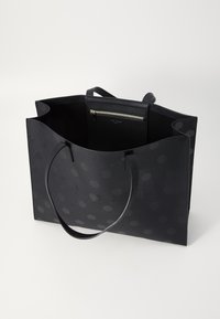 Sac fourre-tout noir avec une surface texturée, présentant des pois en relief et un logo embossé. Poche zippée intérieure pour un rangement sécurisé. Longues poignées.