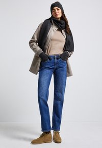 Mujer vestida con jeans azules, chaqueta beige, camisa topo, bufanda gris, guantes y cinta para la cabeza, de pie con las manos en los bolsillos frente a un fondo sencillo.