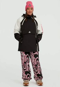 Black and white waterproof jacket with a pink beanie and vibrant patterned pants. Features a triangular logo and elastic cuffs. Brown shoes.