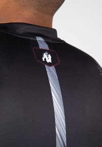 Zwart atletisch shirt met een verticale zilveren streep en een label met een gorillalog op de bovenrug, gladde textuur, getailleerd ontwerp.