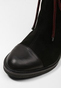 Black ankle boot with a suede upper and a rounded black leather toe cap. Features red and black textured laces and rubber sole.