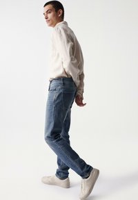Camisa de manga larga de color beige claro combinada con vaqueros de mezclilla azules y zapatillas beige. Los vaqueros tienen un corte relajado, con bolsillos estándar y costuras.