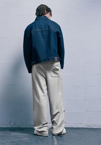 Személy egy falnak háttal, sötétkék farmerdzsekit és világosszürke melegítőnadrágot visel, a hátsó zsebében a "Stranger Things" logójával.