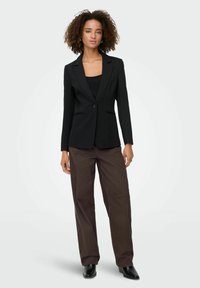 Femme aux cheveux bouclés portant un blazer noir, un haut noir, un pantalon ample marron et des bottines noires, se tenant devant un fond uni.