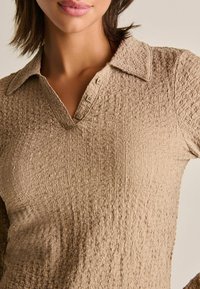 Beige textured långärmad polo med en krage och tre knappar fram. Tyget har en rynkad yta.