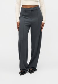 Γκρι παντελόνι με ψηλή μέση και φαρδιά γραμμή, με λεπτομέρειες πιέτας μπροστά και λεία υφή υφάσματος. Φορεμένο με μαύρα μυτερά παπούτσια.