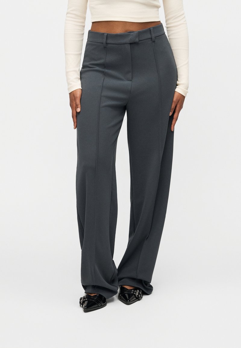 Γκρι παντελόνι με ψηλή μέση και φαρδιά γραμμή, με λεπτομέρειες πιέτας μπροστά και λεία υφή υφάσματος. Φορεμένο με μαύρα μυτερά παπούτσια.