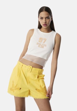 Witte mesh crop top met "97" opdruk, gecombineerd met gele shorts met geborduurde bladeren en ananassen, een franjeszoom en een ceintuur in de taille.