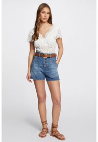 Haut en dentelle blanche à manches courtes, short en denim taille haute avec boutons devant, et ceinture en cuir marron. Sandales à brides marron.