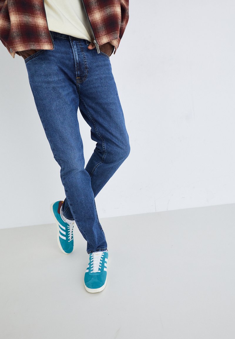 Persona vestida con jeans azules, zapatillas en tonos verdes y blancos, camiseta blanca y chaqueta de cuadros rojos, de pie sobre un fondo liso de suelo y pared blancos.
