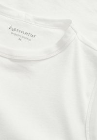 Vit ekologisk bomullstopp med rundad halsringning, mjukt material och etikett med varumärket och storlek som anger hållbart material.