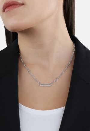 Collana in argento con link allungati e un pendente a forma di spilla di sicurezza indossata al collo di una donna con un blazer nero e una maglietta bianca.